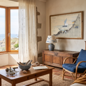Coin de salon lumineux de style bord de mer avec un fauteuil en rotin bleu, une table basse en bois ornée de coquillages et un grand cadre en bois affichant une carte marine vintage au-dessus d'un coffre