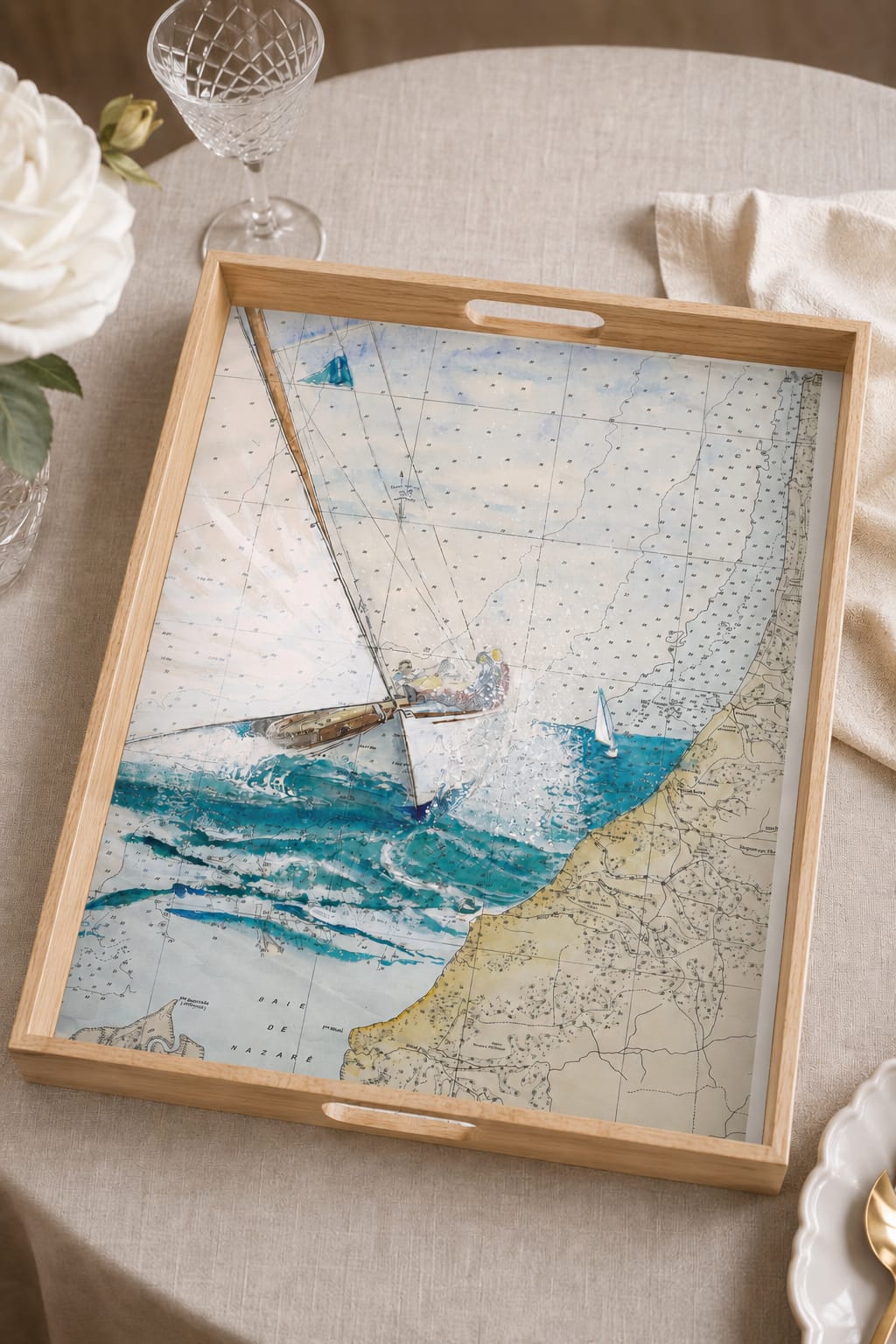 Plateau en bois présentant une aquarelle de voilier naviguant sur une carte marine ancienne, posé sur une table dressée.
